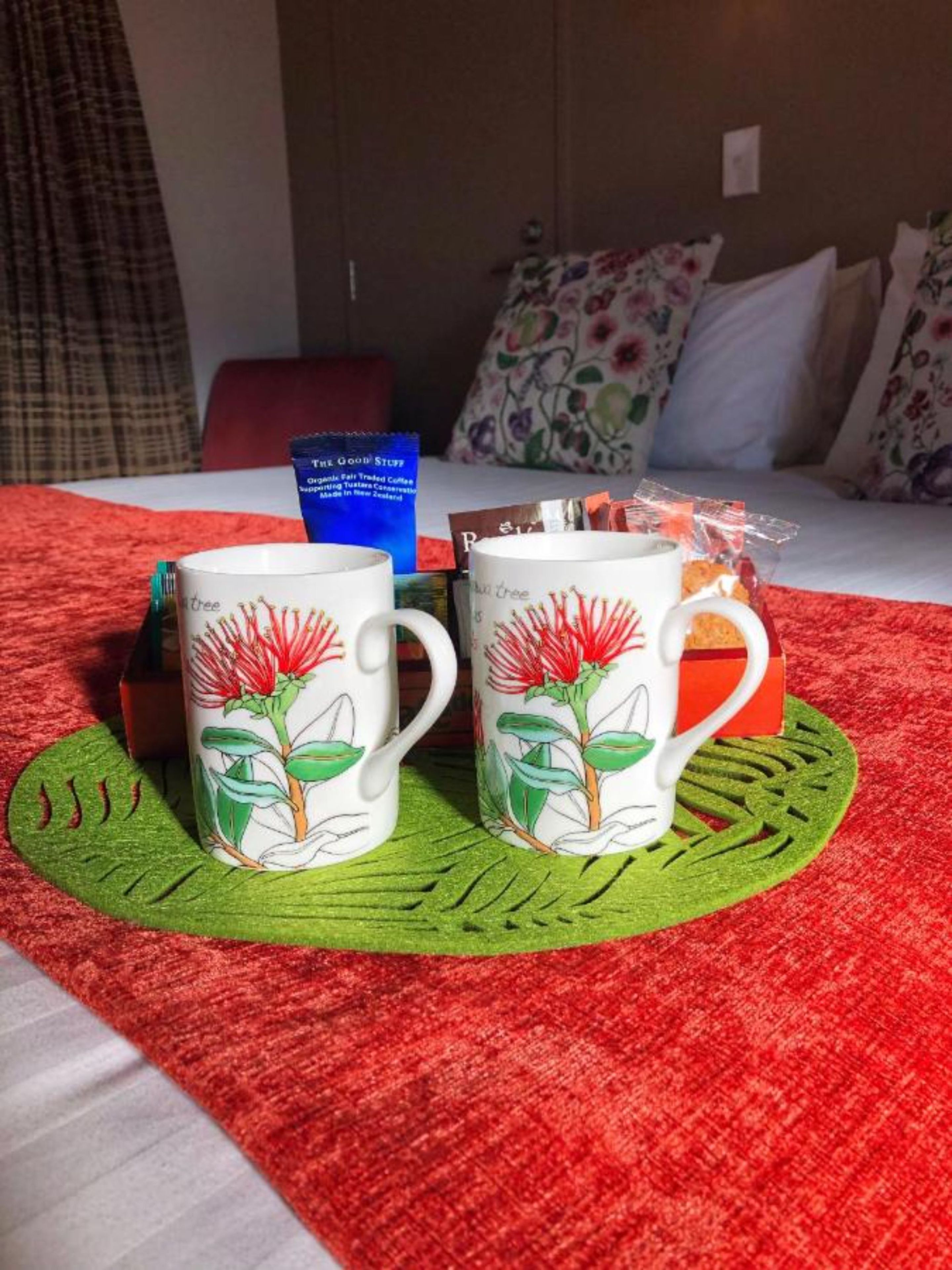 Two white mugs with red flowers on a green placemat on a bed with a red blanket.