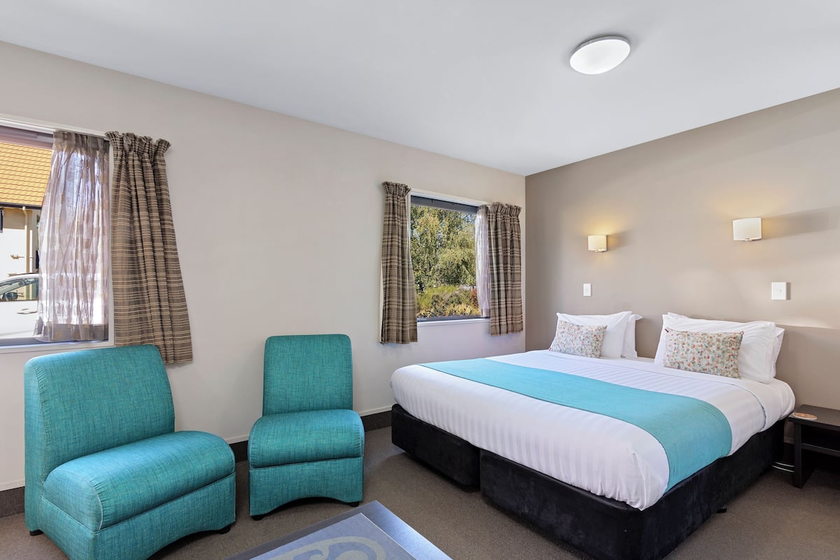 Una habitación de hotel con una cama grande, dos sillones a juego, un ventanal con cortinas y luces fijadas a la pared.