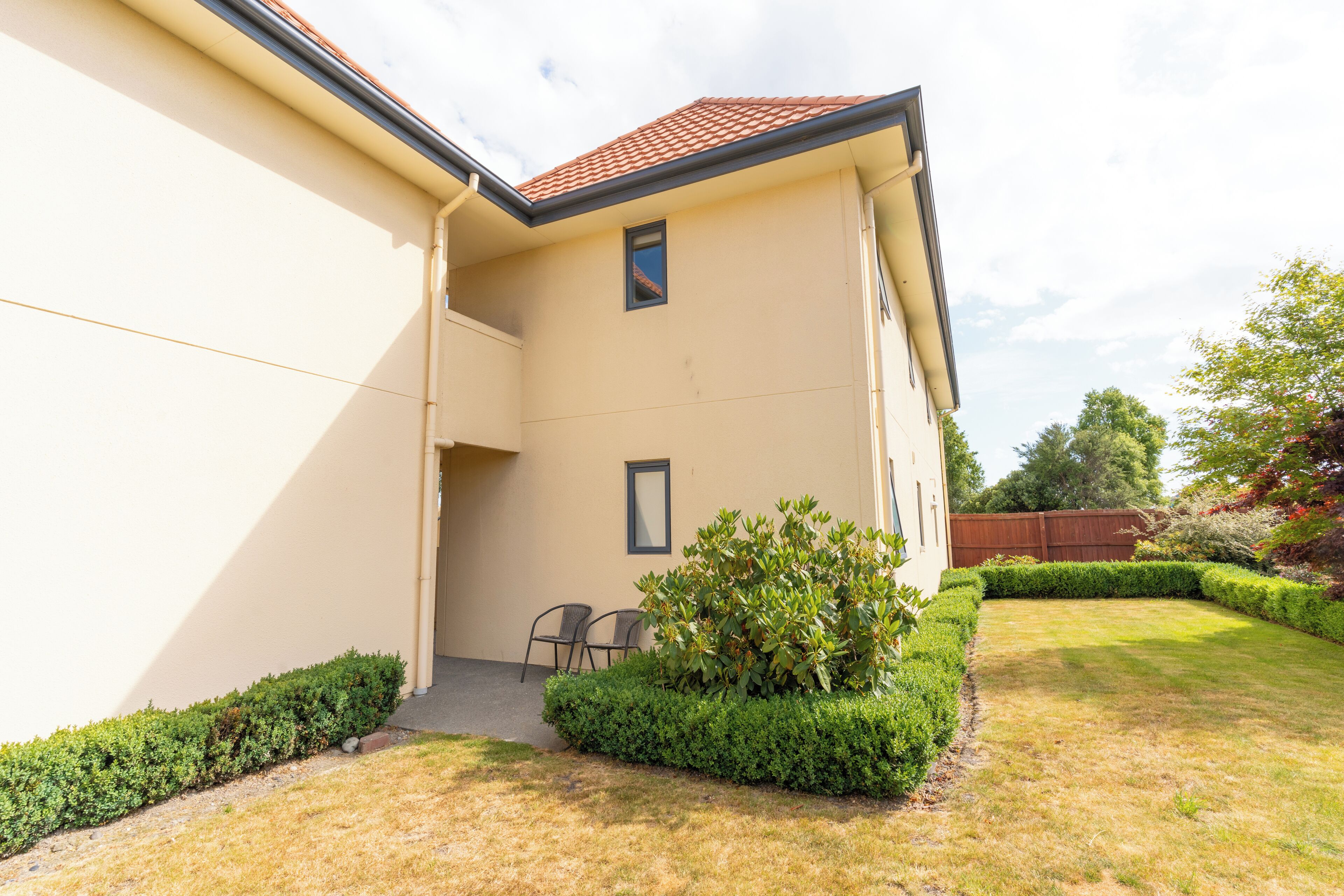 Ein Haus mit heller Fassade, einem roten Ziegeldach und einem kleinen Fenster. Vor dem Haus liegt eine gepflegte Rasenfläche mit geschnittenen Büschen und im Hintergrund ein Holzzaun.