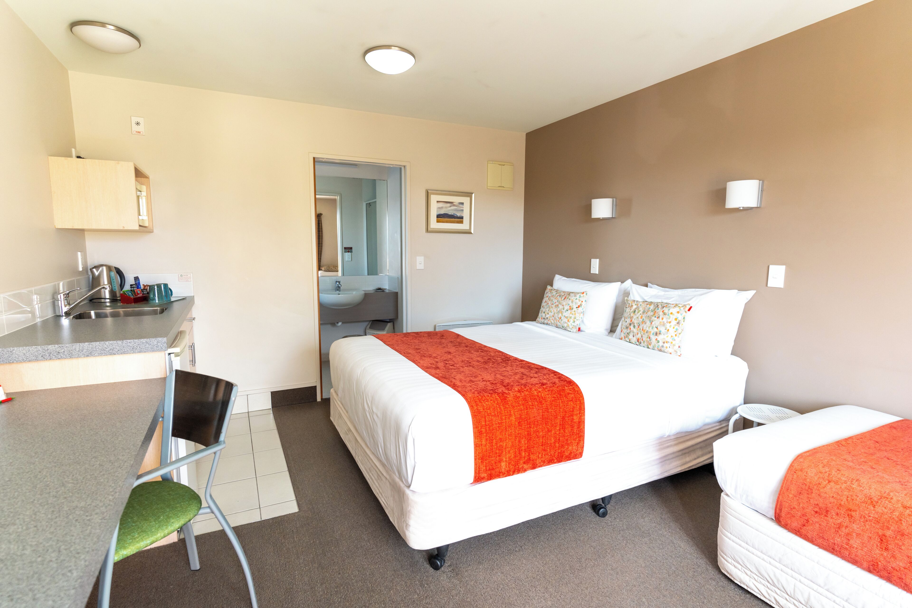 A hotel room with a bed, a desk with a chair, a kitchenette, and a bathroom visible through an open door.