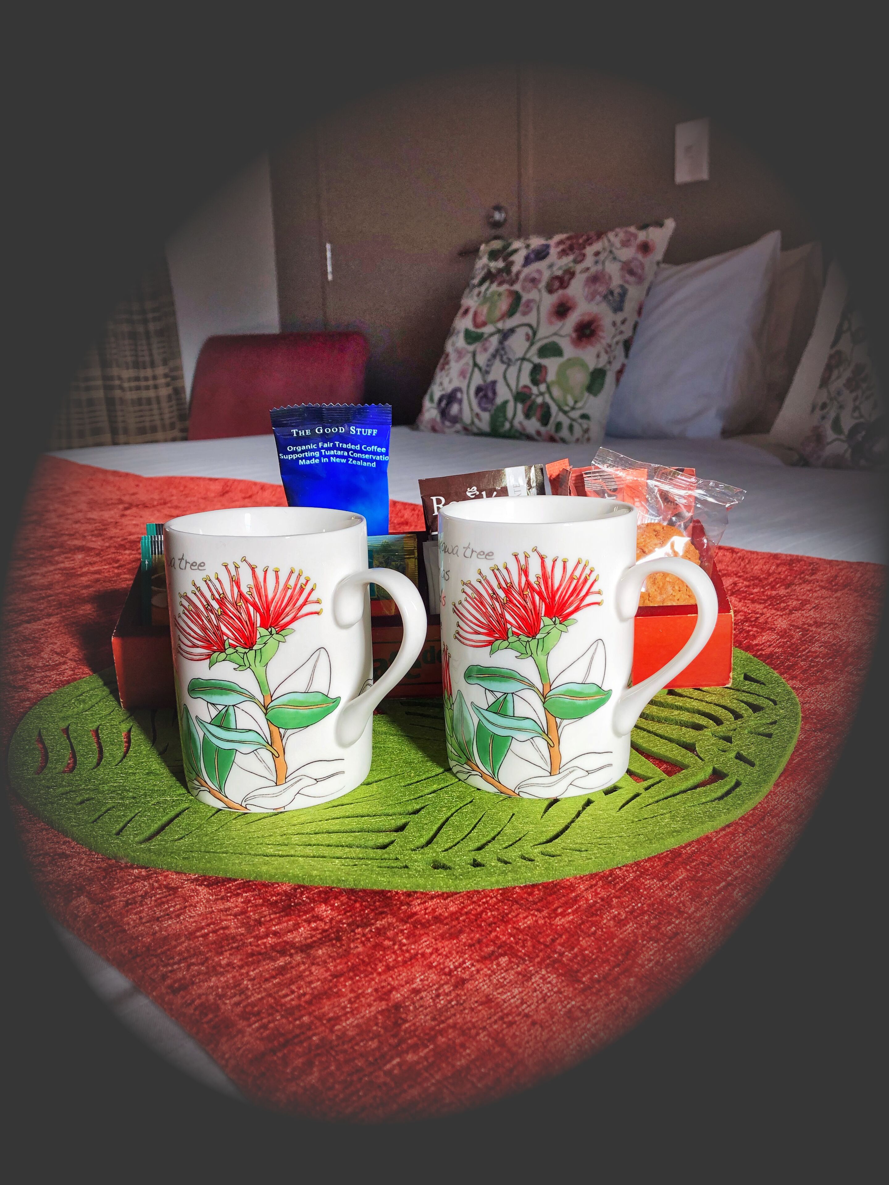 Two white mugs with floral designs on a red textured tablecloth, a blue packet, and a red box on a bed with a red blanket and floral pillows.
