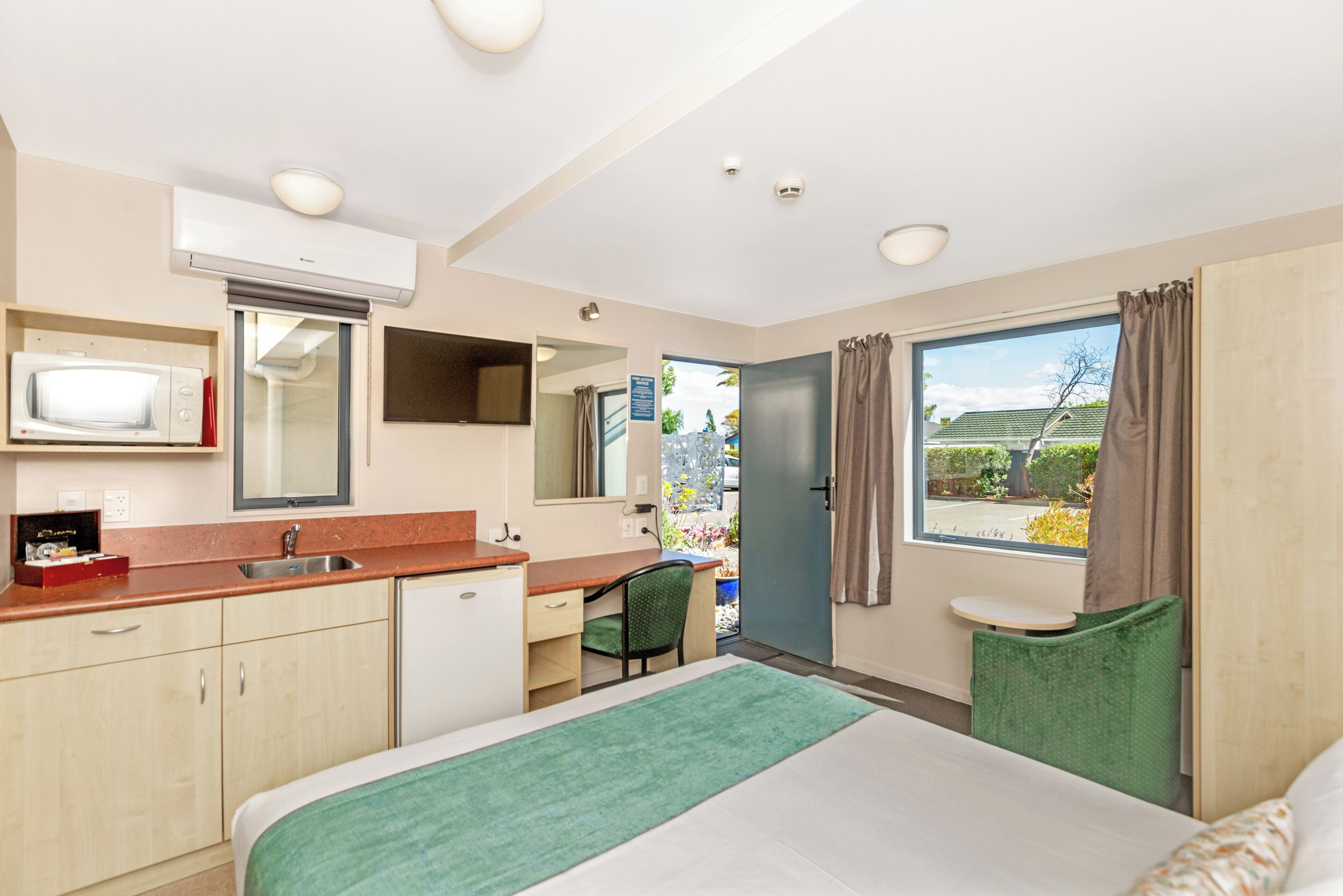 A hotel room with a bed, a desk, a chair, a small table, a window with curtains, a microwave, a sink, and an air conditioning unit.