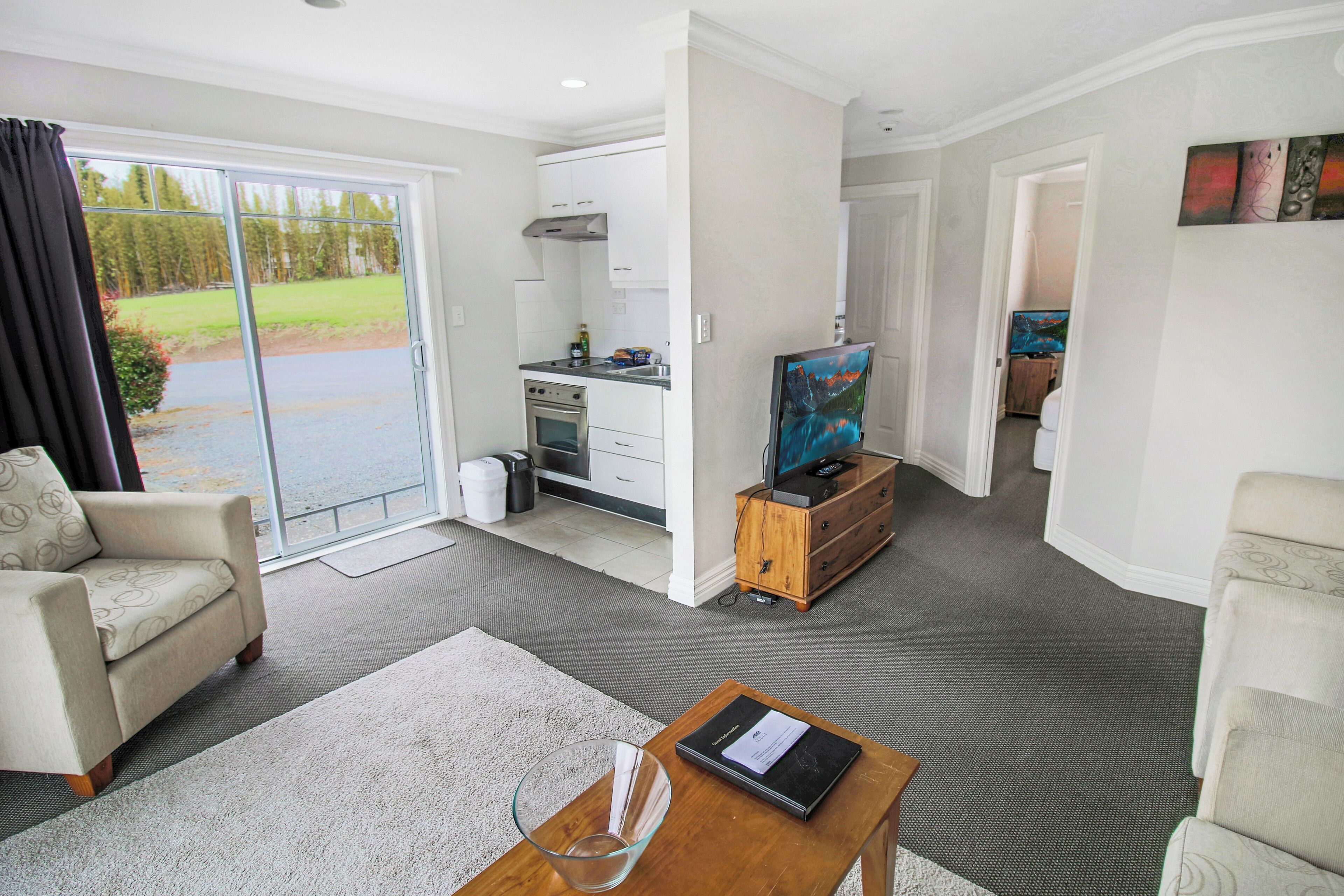 A modern living room with a sofa, a coffee table, and a television. There is a sliding glass door leading to a balcony with a view of trees.