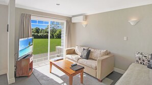 Una sala de estar con televisor, sofá, mesa de centro y vista a un paisaje verde a través de una puerta corrediza.