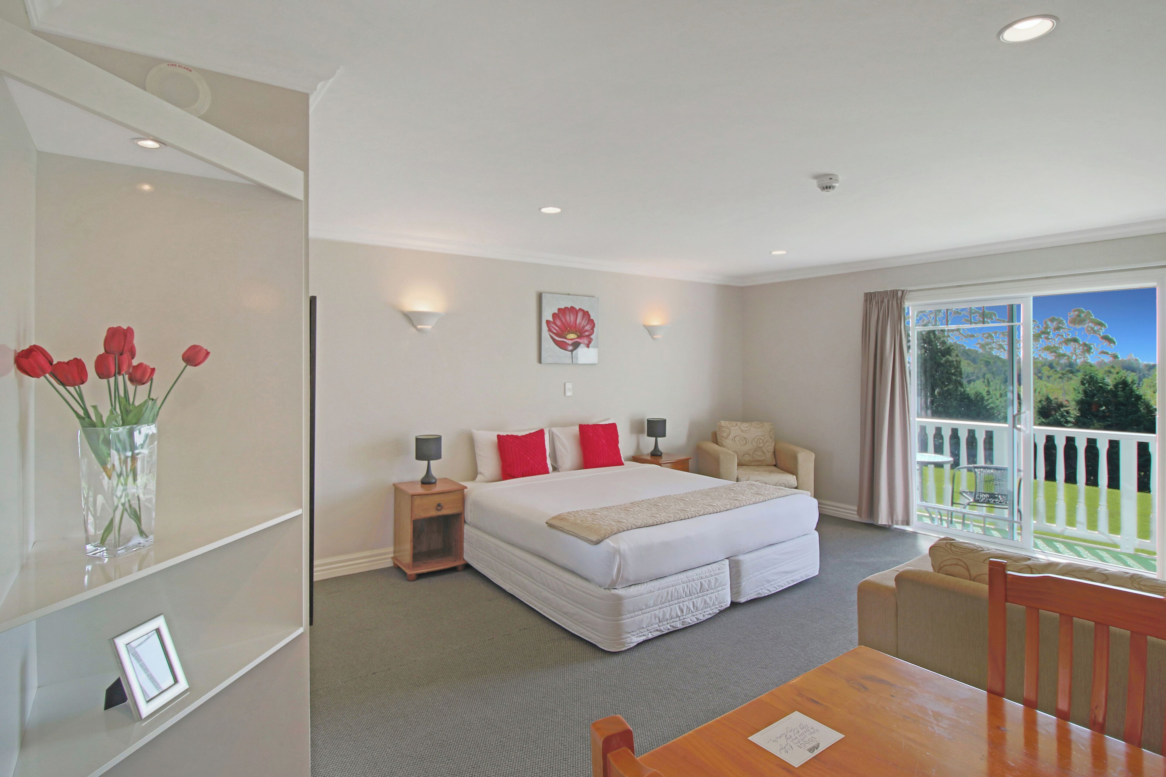 A hotel room with a large bed, bedside tables, a chair, a small table, a window with a view of the outdoors, and a vase with flowers on a shelf.