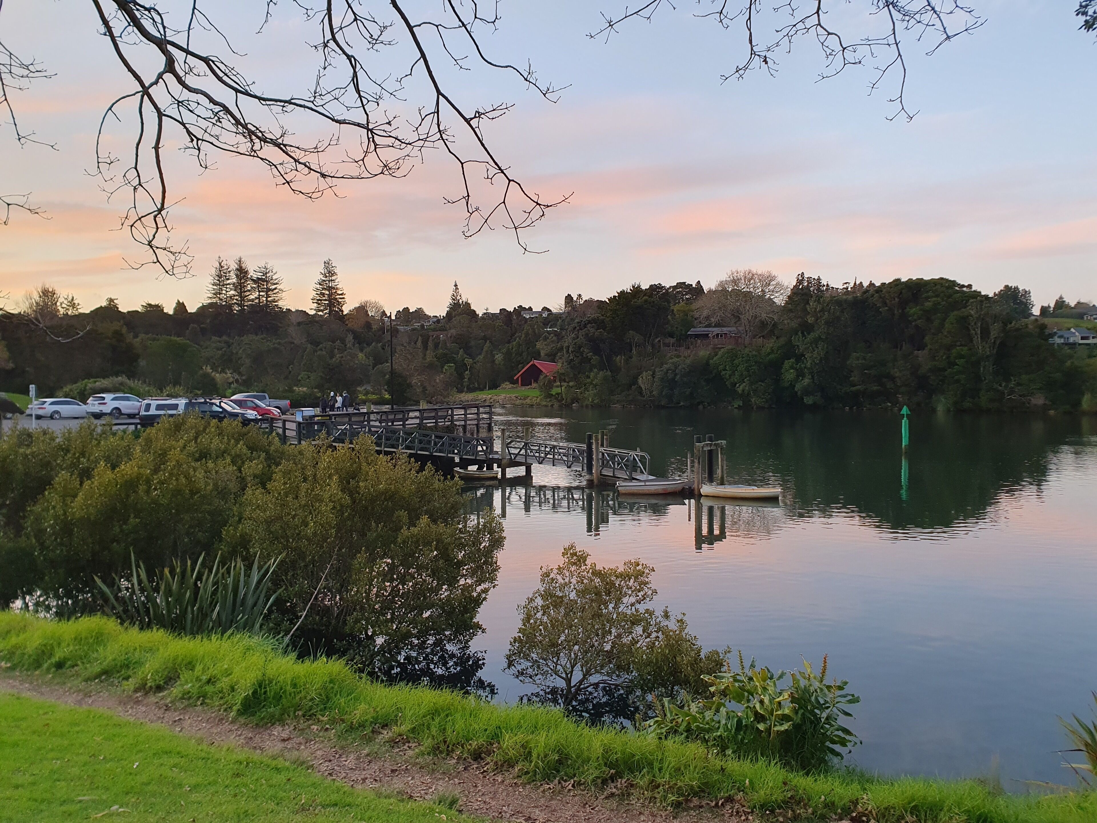 A lakeside view with a dock, parked cars, and a red building in the background.