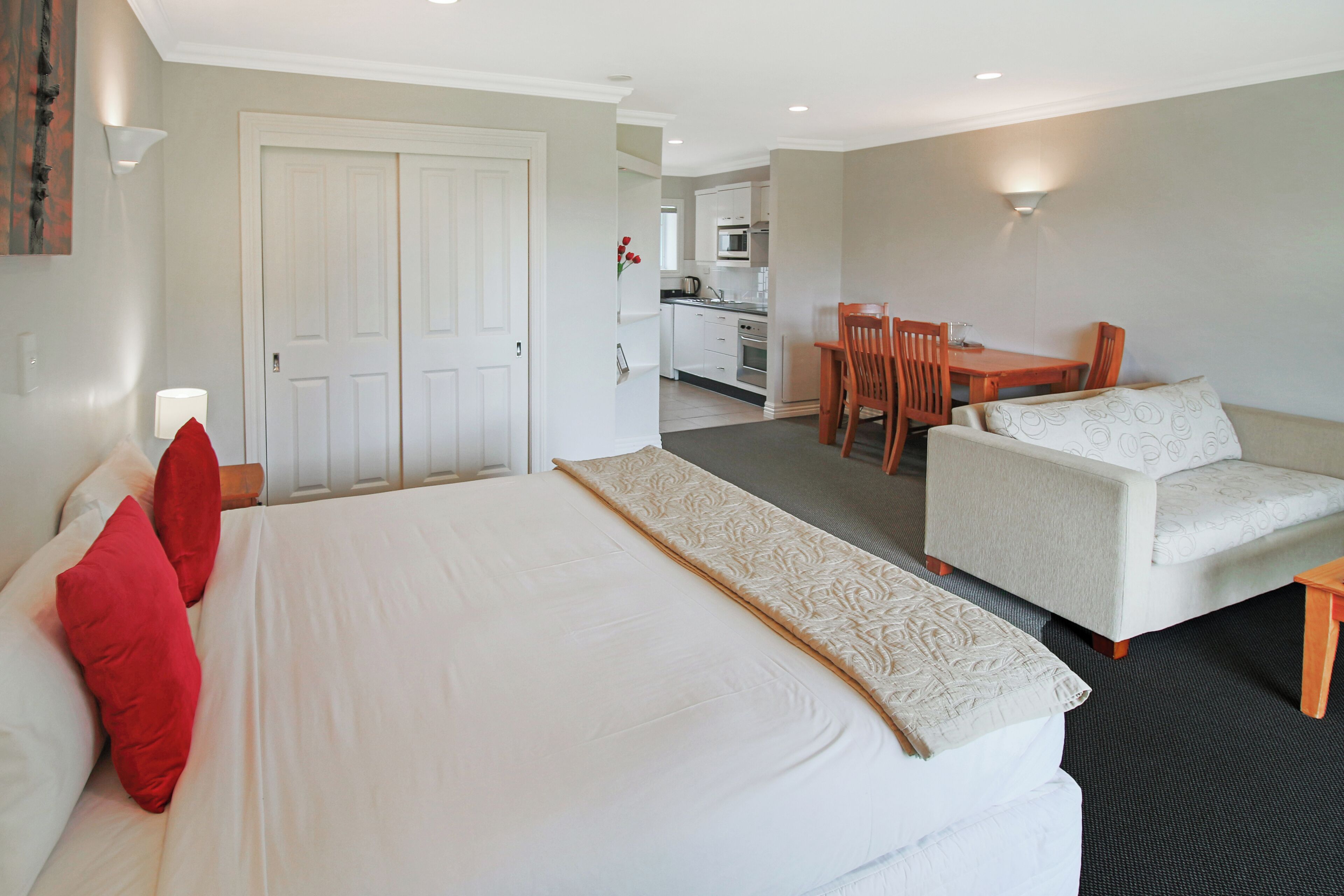 A hotel room with a bed, a sofa, a dining table with chairs, a kitchen area, and a door.