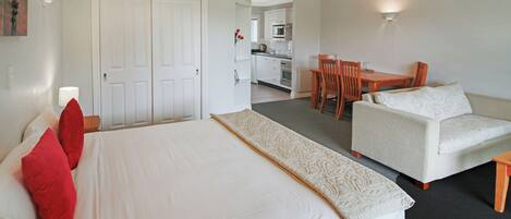 A hotel room with a bed, a sofa, a dining table with chairs, a kitchen area, and a door.