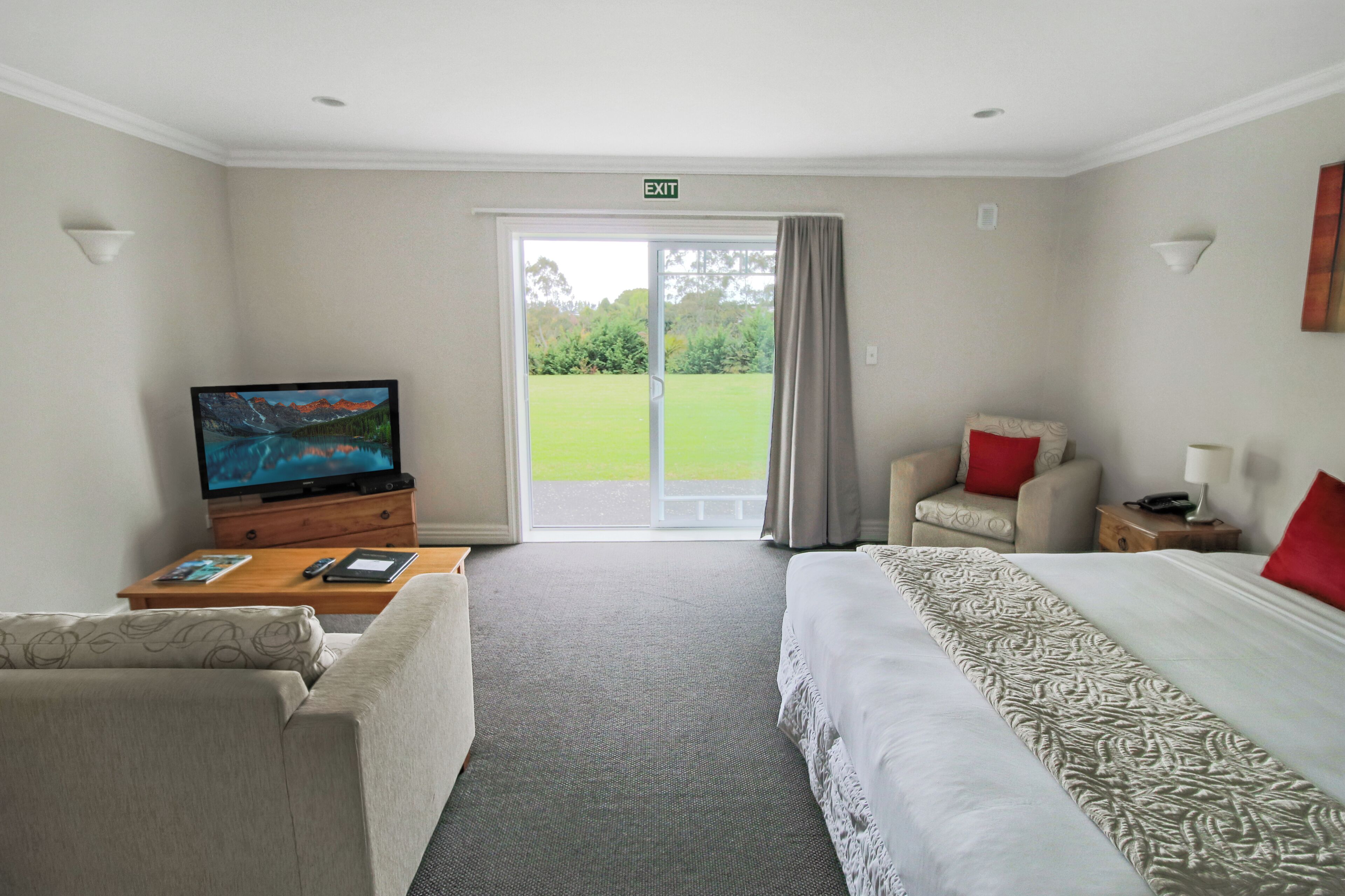 A hotel room with a bed, a sofa, a TV, and a view of a green landscape through the door.