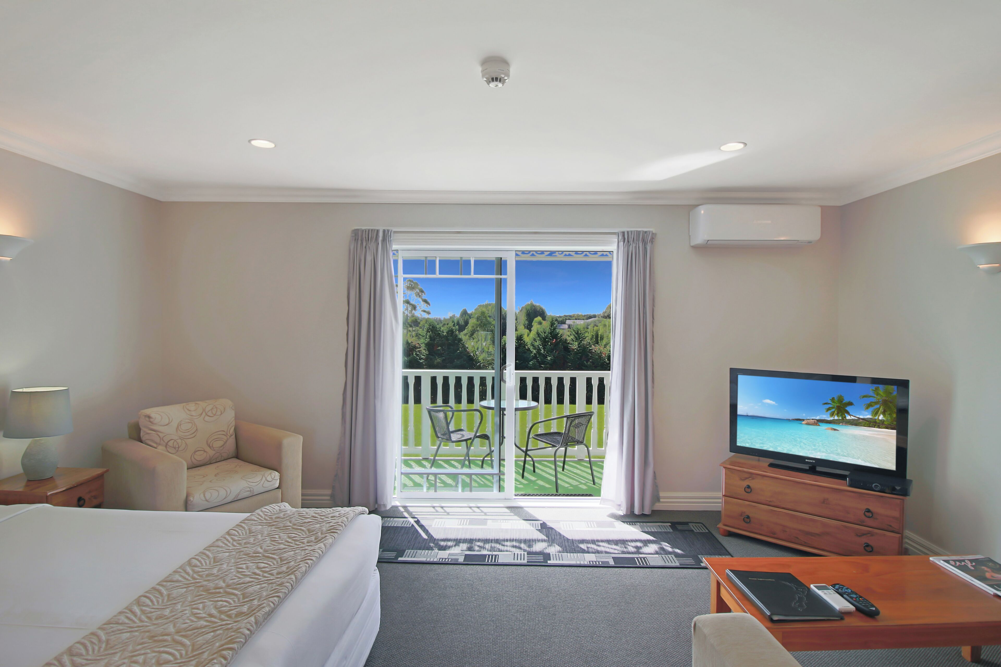 A hotel room with a bed, a TV on a wooden stand, a desk with a remote control, and a balcony with chairs and a view of the sea.