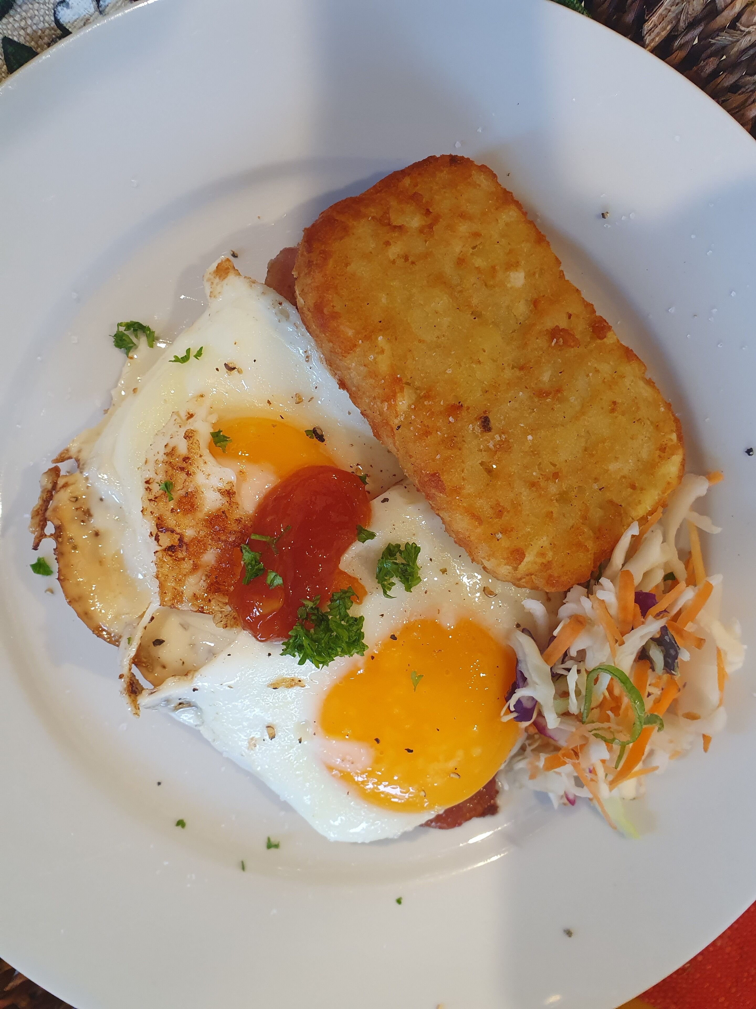 A plate with two fried eggs, a slice of hash brown, and a side of coleslaw.