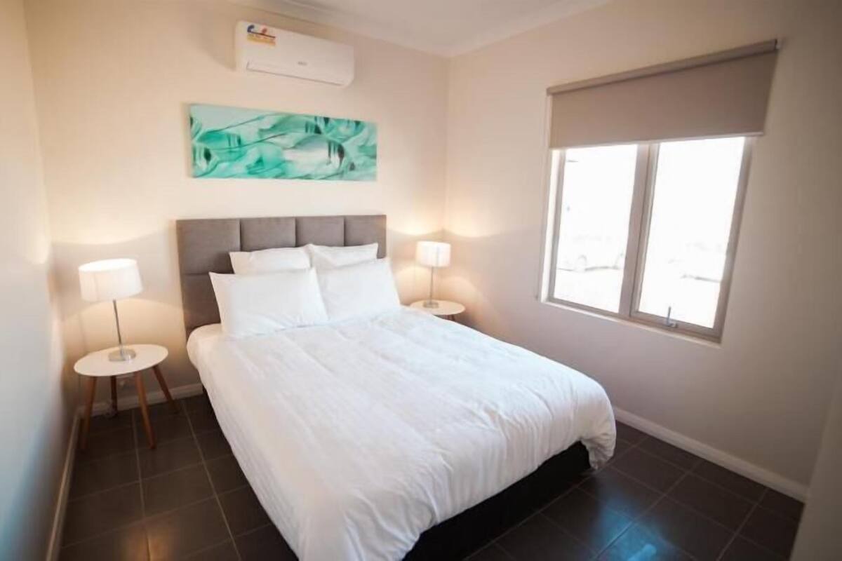 Un dormitorio con una cama, dos mesitas de noche, aire acondicionado instalado en la pared, una ventana con persiana y un cuadro en la pared.