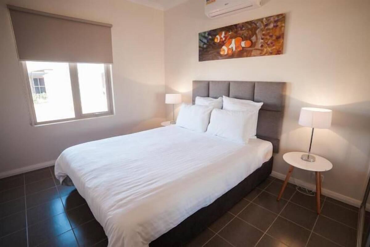 Un dormitorio con cama, dos lámparas de noche, una ventana con cortina y un cuadro en la pared.