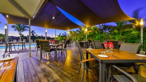 An outdoor dining area with wooden tables and chairs, a pool, and palm trees in the background. - Exmouth Escape Resort (Exmouth)