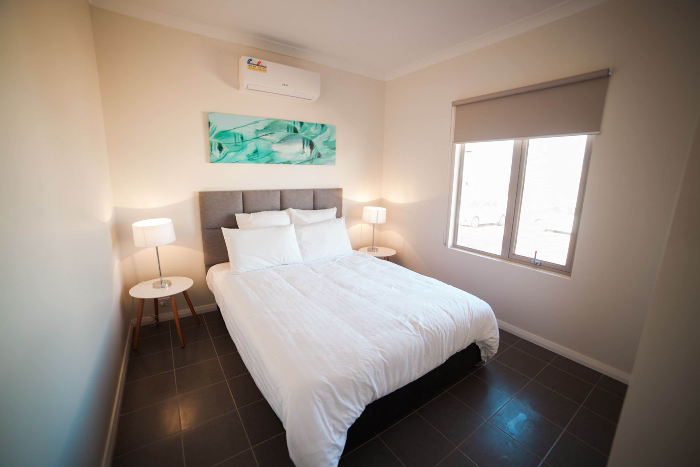 A neatly arranged bedroom with a large bed, two bedside tables with lamps, a ceiling air conditioner, a window with a blind, and a wall-mounted artwork.