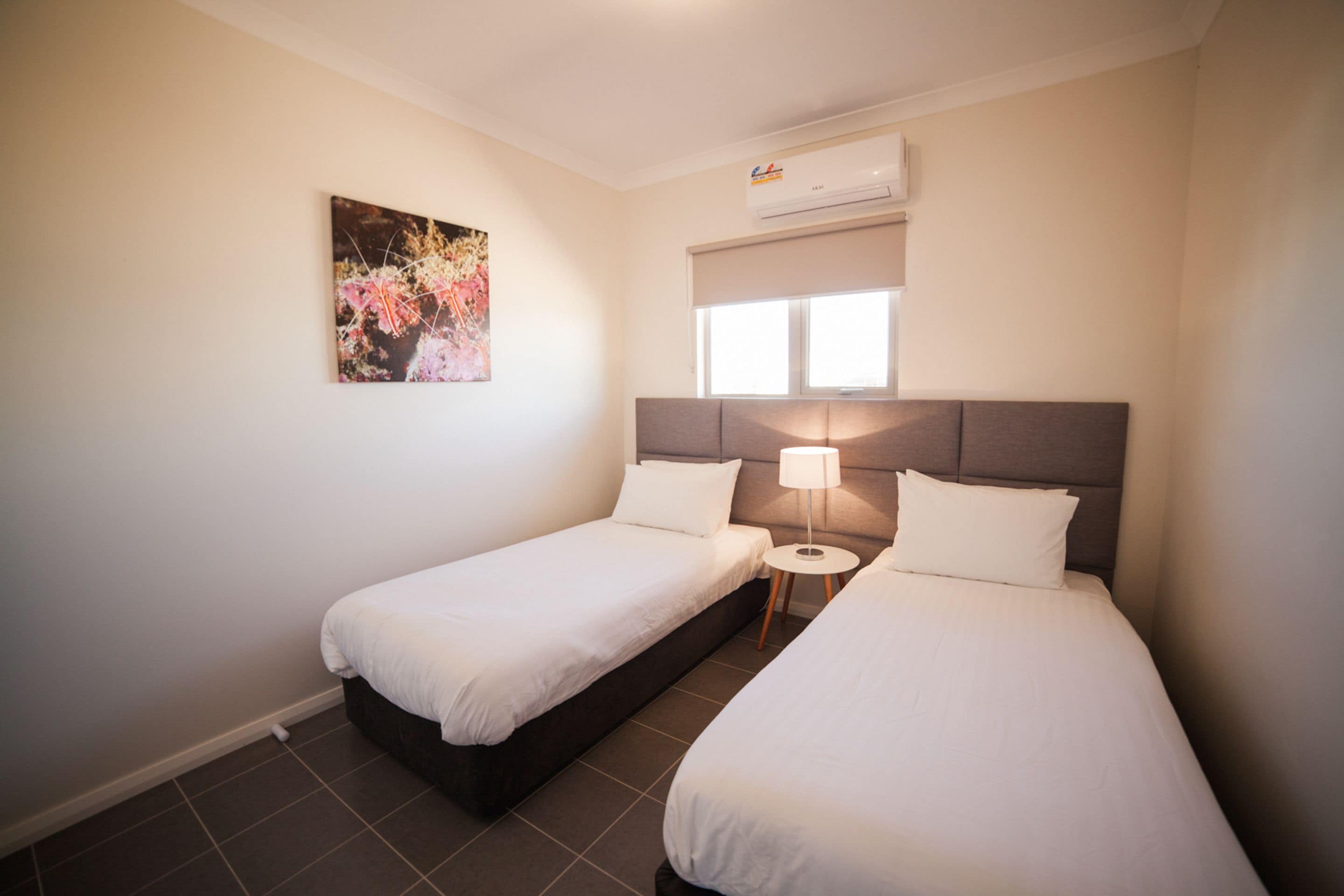 A hotel room with two single beds, a small round table, a wall-mounted air conditioner, and a window with blinds.