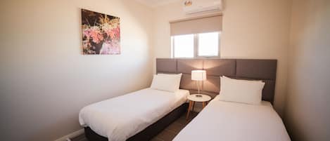A hotel room with two single beds, a small round table, a wall-mounted air conditioner, and a window with blinds.