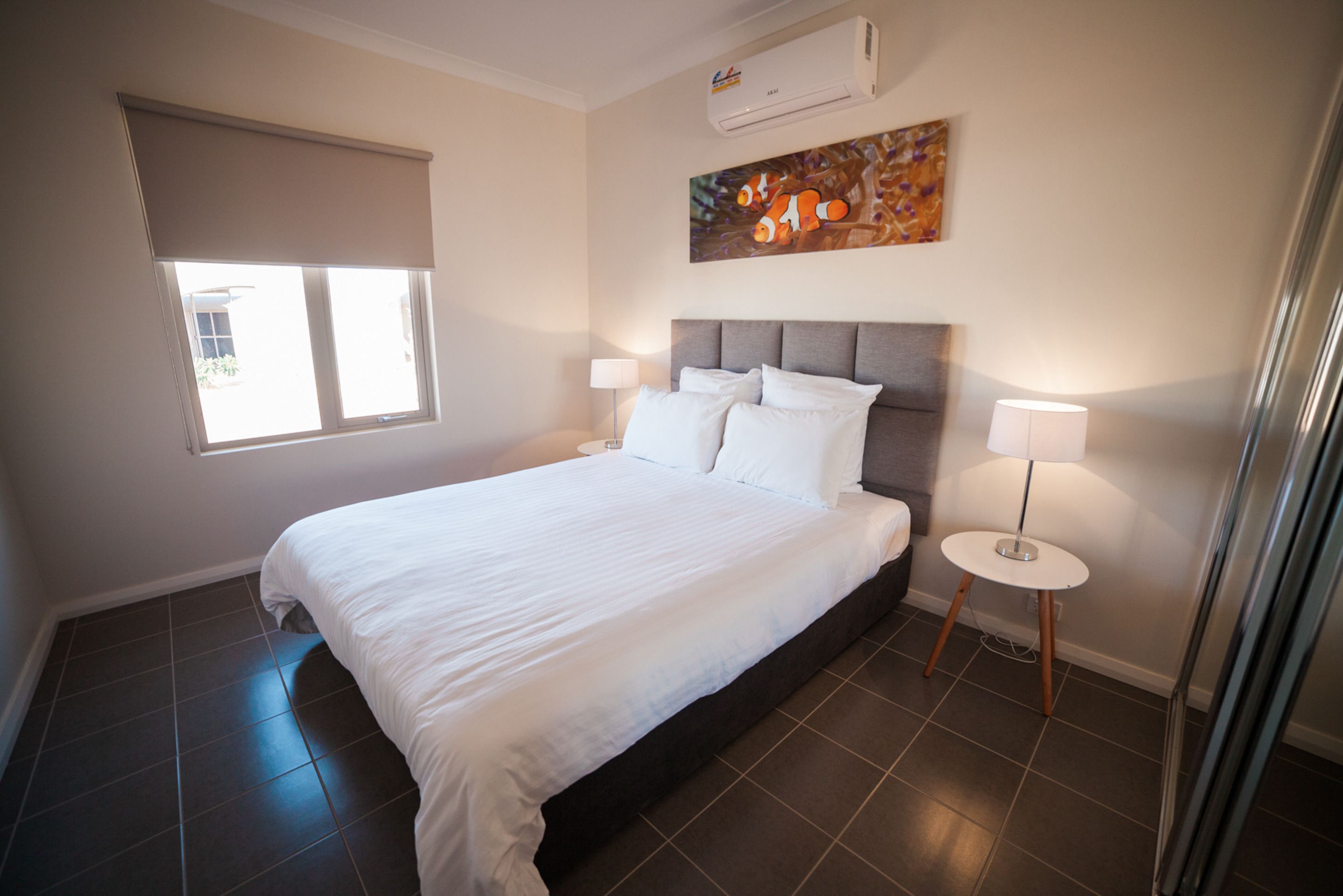 Une chambre avec un grand lit, deux tables de chevet avec des lampes, une fenêtre avec un store et un appareil de climatisation fixé au mur.