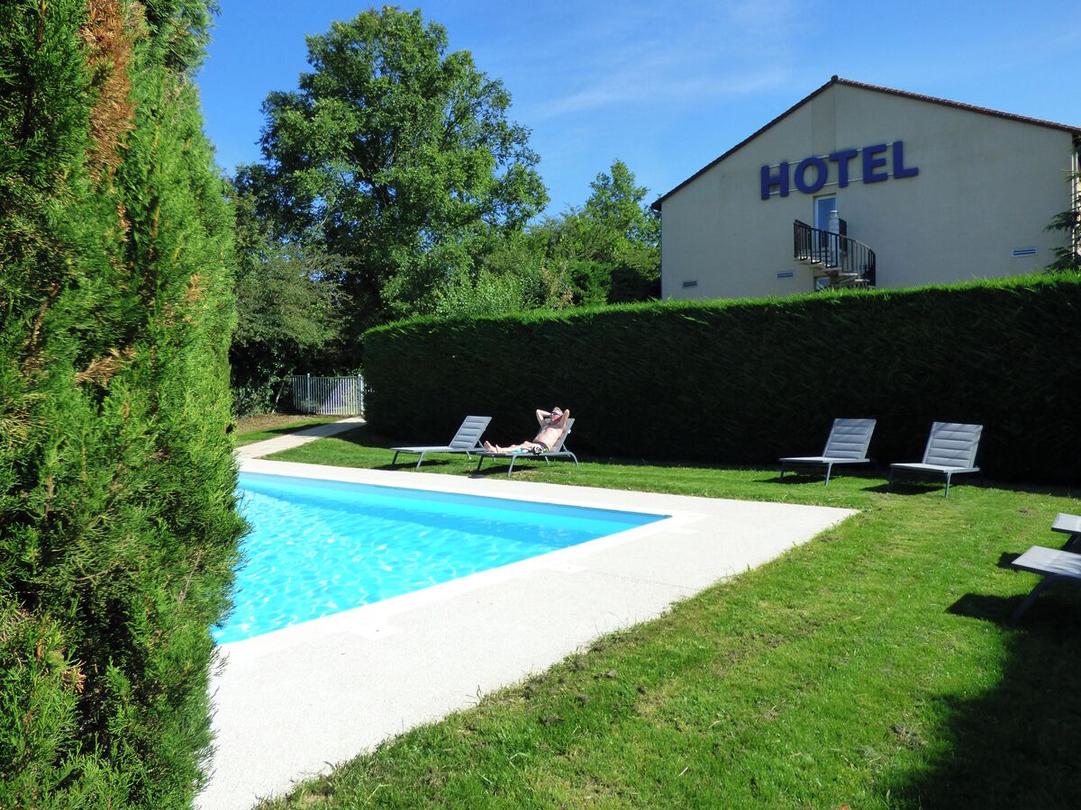 A hotel with a swimming pool, lounge chairs, and a hedge.