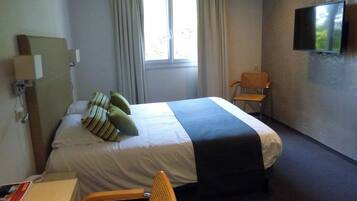 Ein Hotelzimmer mit Bett, Schreibtisch, Stuhl, Fernseher und einem Fenster mit Vorhängen.
