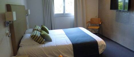 Habitación de hotel con cama, escritorio, silla, televisor y ventana con cortinas.