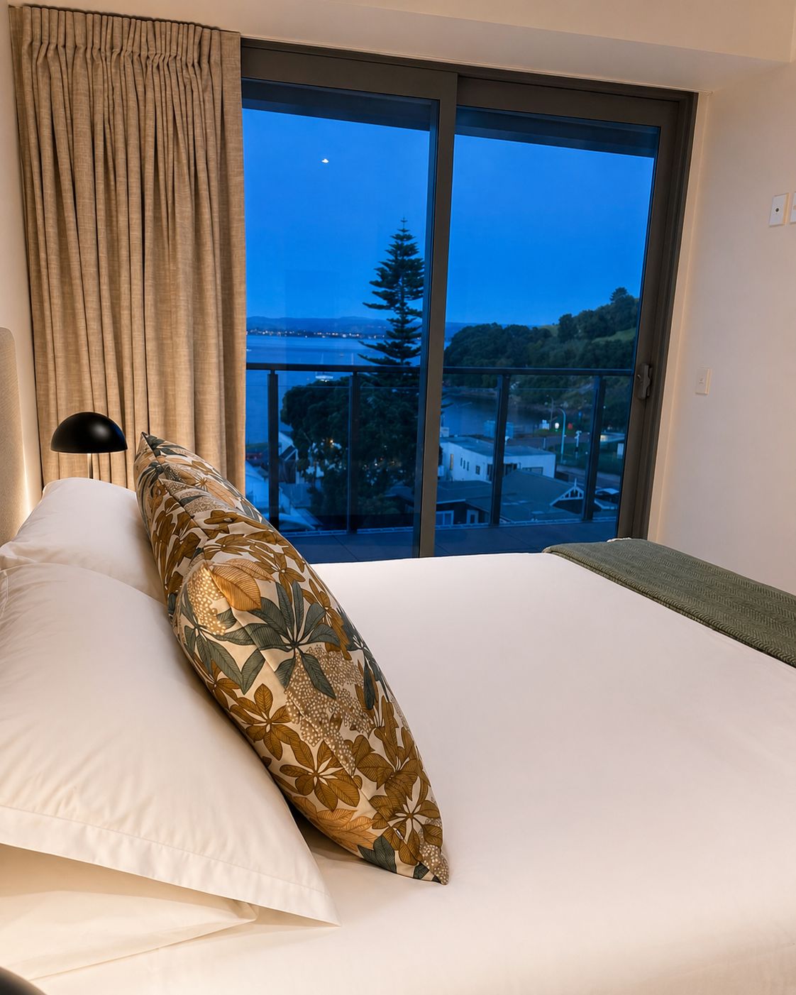 A hotel room with a bed, a nightstand, a lamp, and a large window with a view of a body of water and trees.