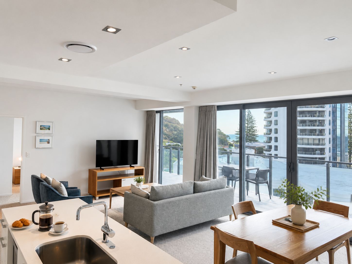 Ein modernes Wohnzimmer mit Flachbildfernseher, grauem Sofa, Essbereich und einem Balkon mit Blick auf Gebäude und Hügel.