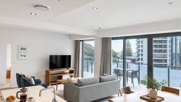 Ein modernes Wohnzimmer mit Flachbildfernseher, grauem Sofa, Essbereich und einem Balkon mit Blick auf Gebäude und Hügel.
