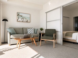 A modern living room with a sofa, a coffee table, a laptop, a chair, and a bed with a sliding door. - The Pacific Apartments (Tauranga)
