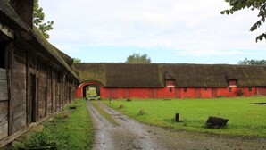 Una construcción rural con techo de paja y paredes rojas, un camino de grava que conduce a un arco, y un campo verde en primer plano.