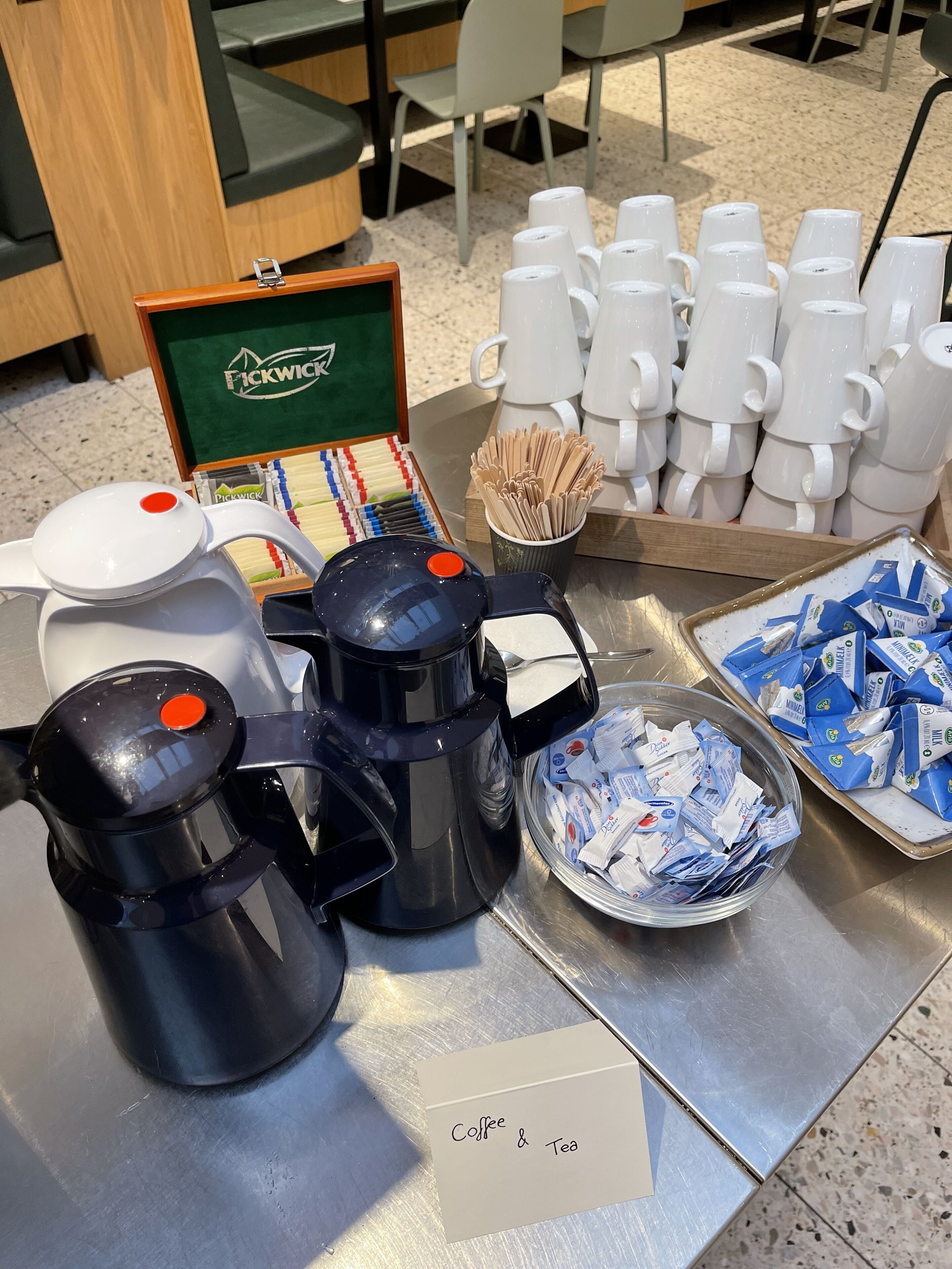 Estación de café y té con varias máquinas de café, vasos desechables y un recipiente con sobres de café.
