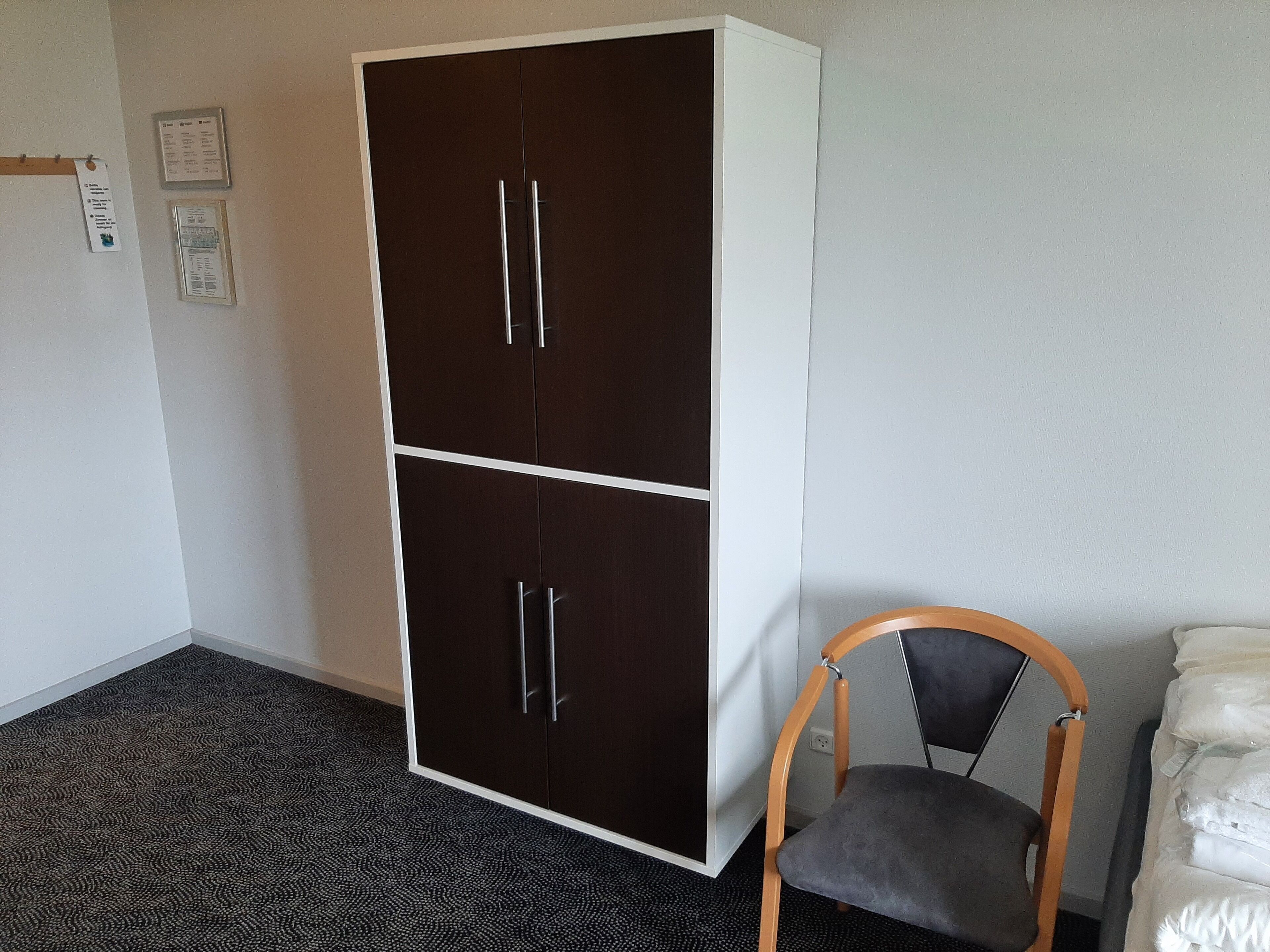 Ein Zimmer mit einem dunkelbraunen Kleiderschrank, einem Holzstuhl und einem Bett mit weißer Bettwäsche.