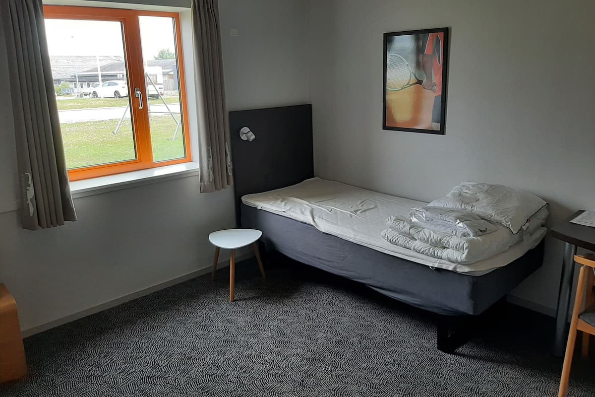 Une petite chambre avec un lit simple, une fenêtre, une tête de lit, un petit tabouret et un tableau accroché au mur.