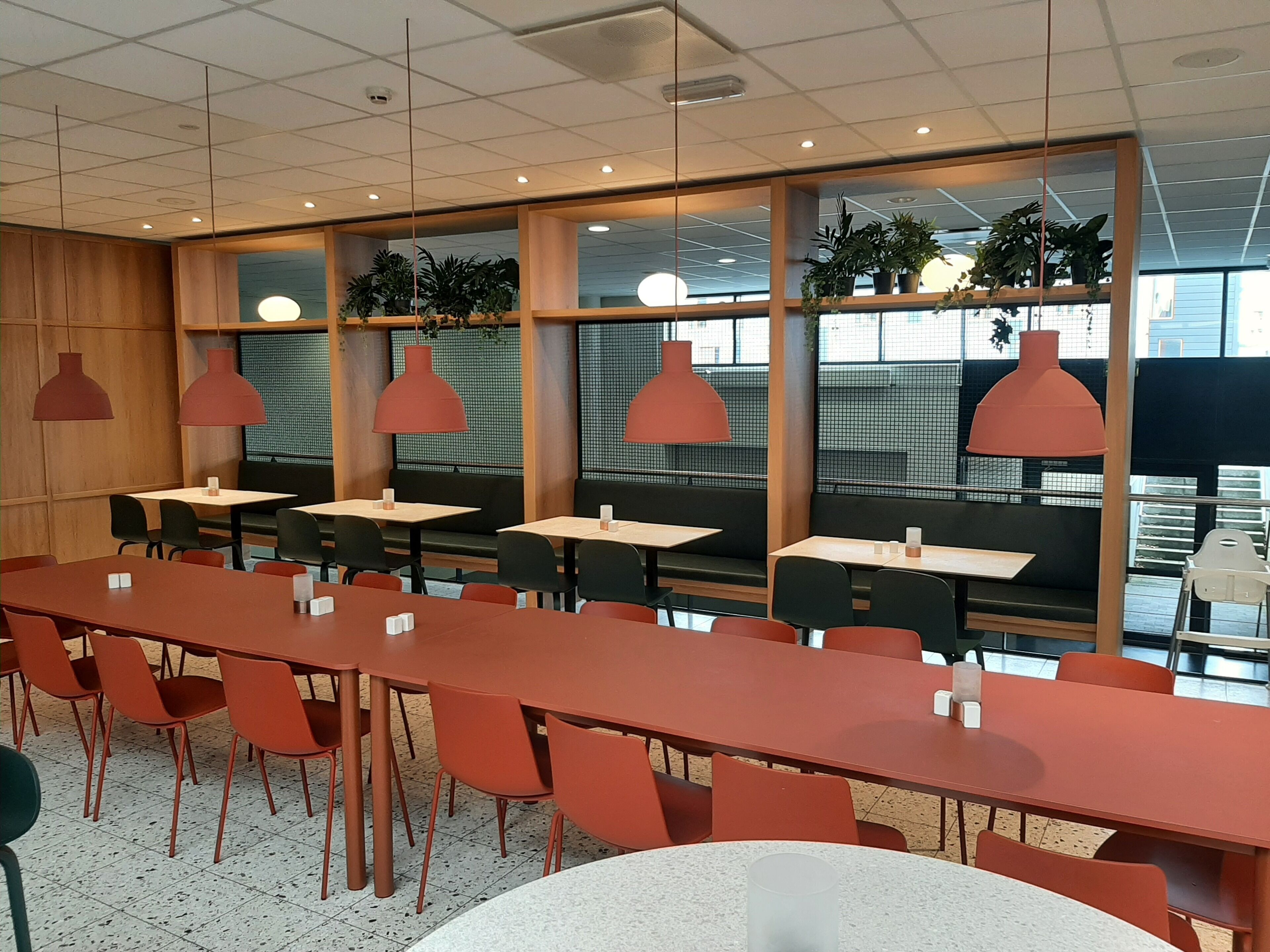 A modern indoor dining area with long red tables and black chairs, wooden paneling, and red pendant lights.