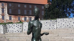 Una estatua de bronce de una figura masculina sobre un pedestal con una placa, ubicada en una zona empedrada con escalones que conducen a un edificio de fachada con tejas.