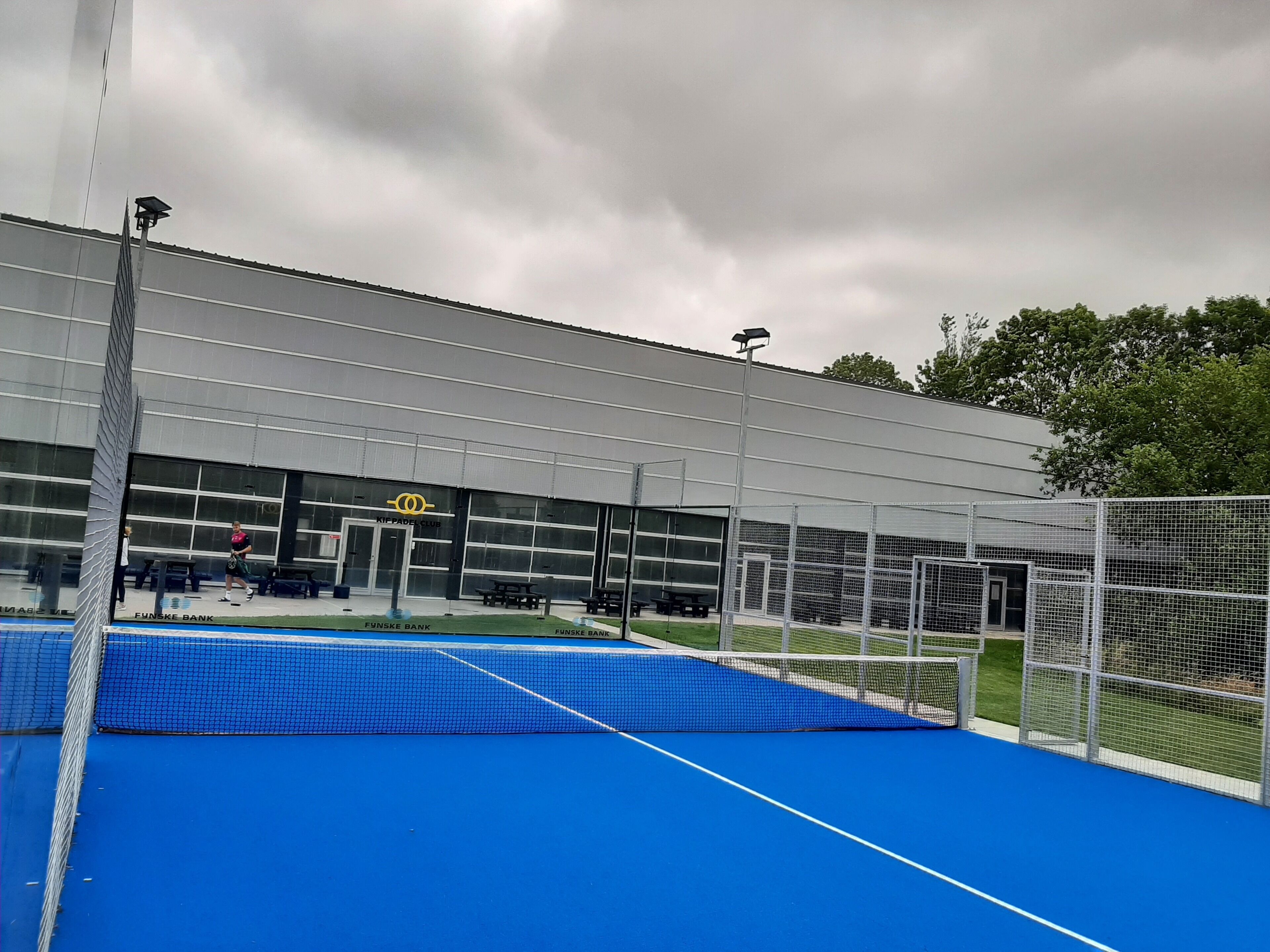 An indoor tennis court with a blue surface, a net, and a building with a sign that reads "KIF PADEL CLUB".