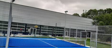 An indoor tennis court with a blue surface, a net, and a building with a sign that reads "KIF PADEL CLUB".