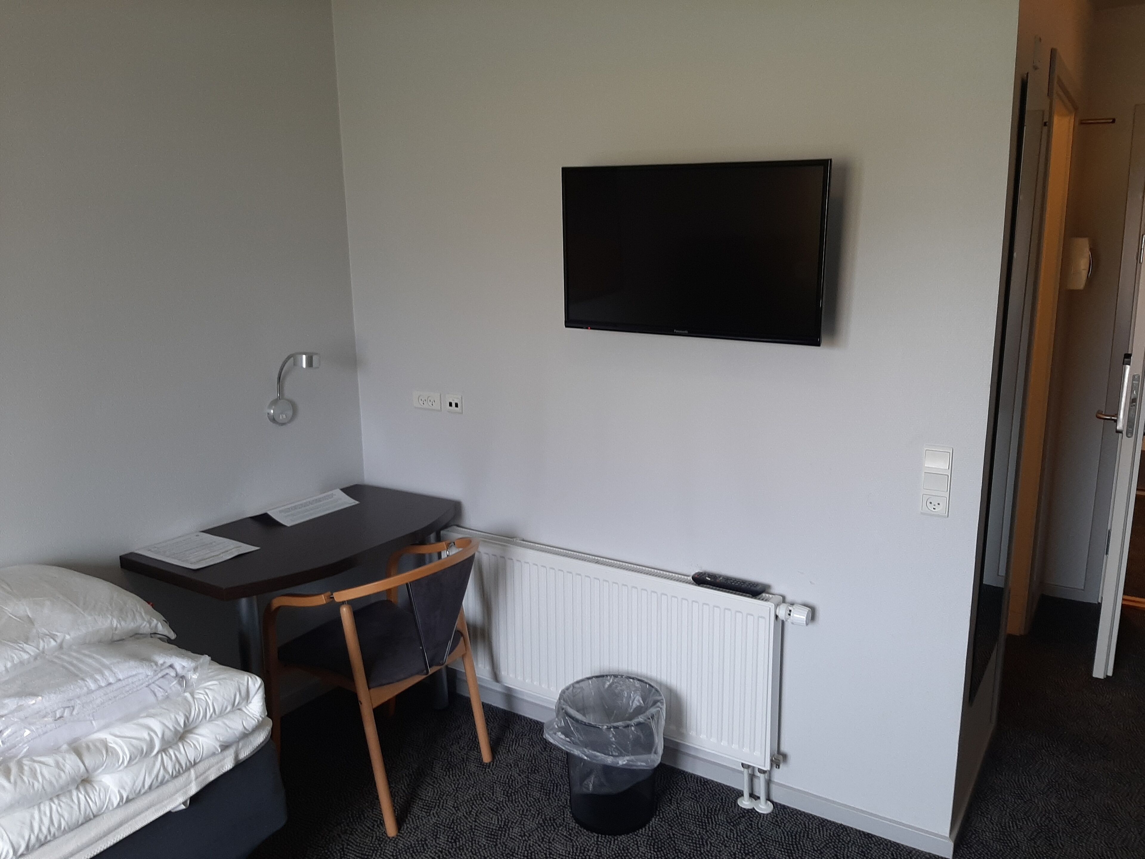 A small hotel room with a bed, a desk, a chair, a television mounted on the wall, a radiator, and a trash bin.