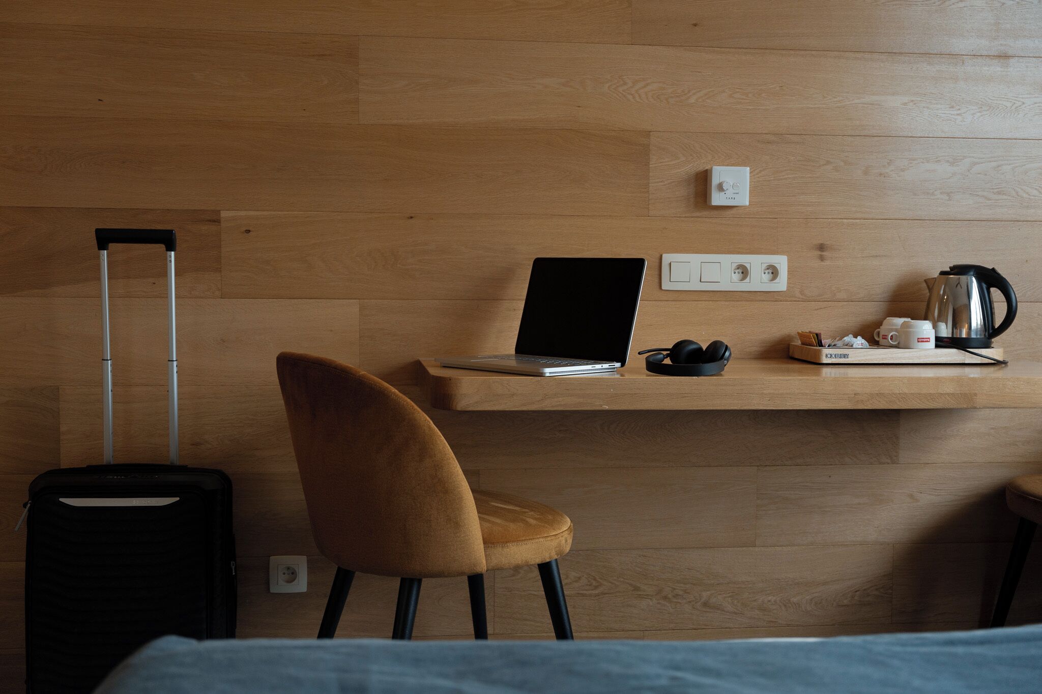 Habitación de hotel con piso de madera, un escritorio con una computadora portátil, una silla, una maleta y una cafetera.