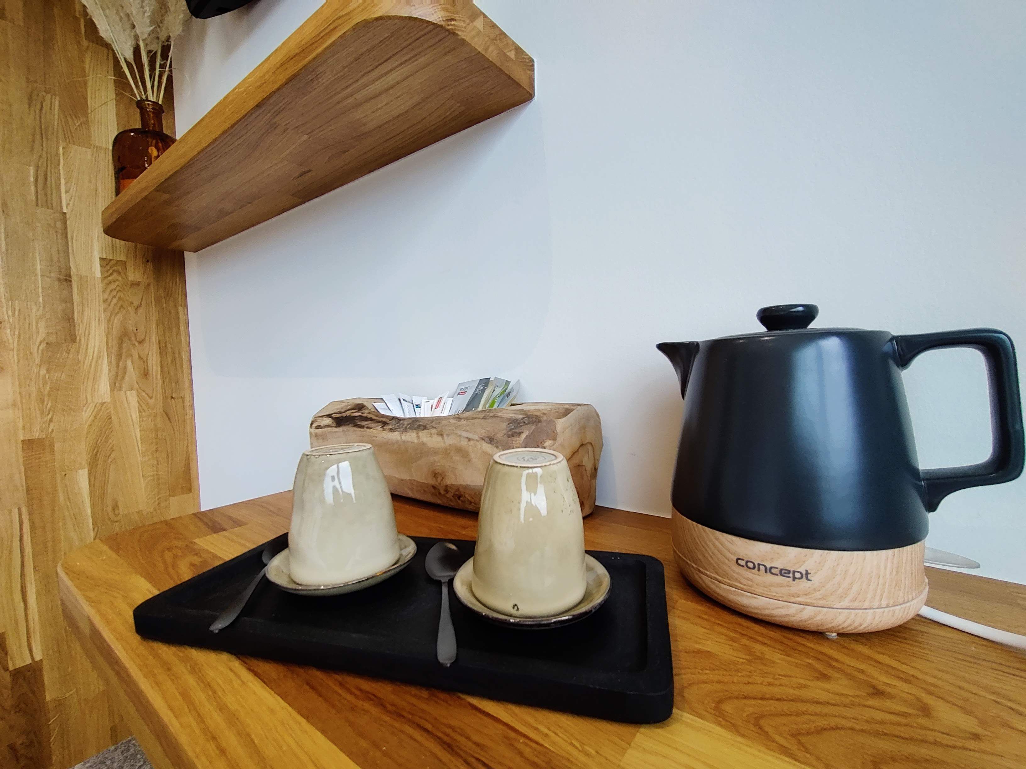 A wooden table with two white ceramic cups, a black spoon, and a black and beige kettle.