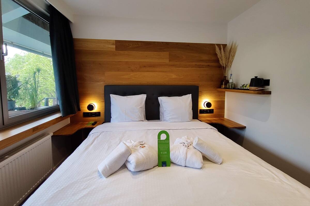 Un lit bien fait, avec du linge de lit et des serviettes blanches, une tête de lit en bois, des lampes de chevet, une fenêtre avec des rideaux et une petite étagère avec quelques objets.