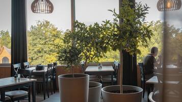 A restaurant with tables and chairs, potted plants, and a view of trees and buildings.