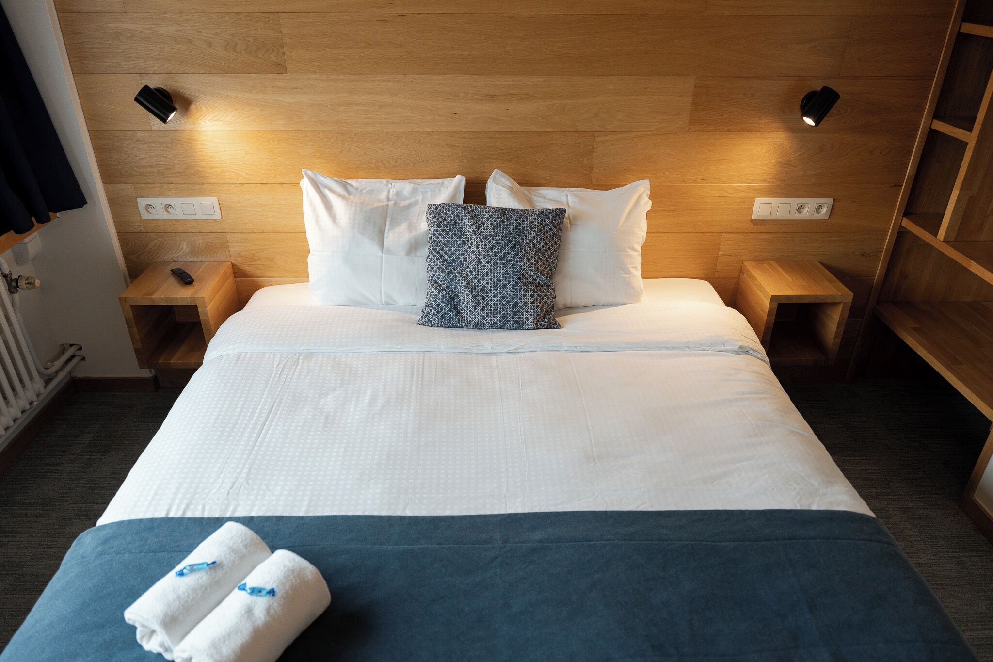Habitación de hotel con cama, dos mesitas de noche, lámpara de pared y toma de corriente.
