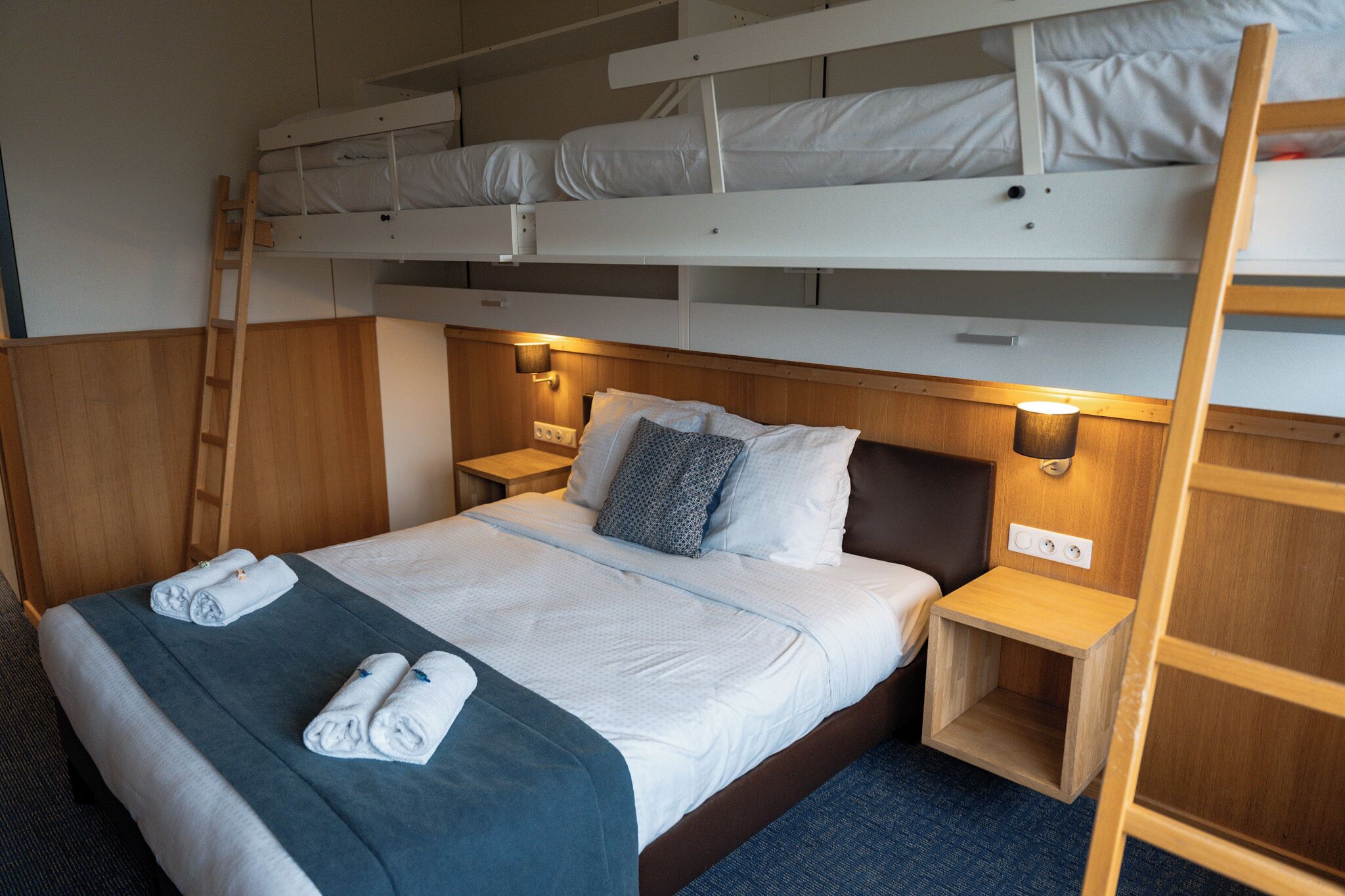A bunk bed room with a wooden ladder, bedside lamps, and neatly folded towels on the bed.