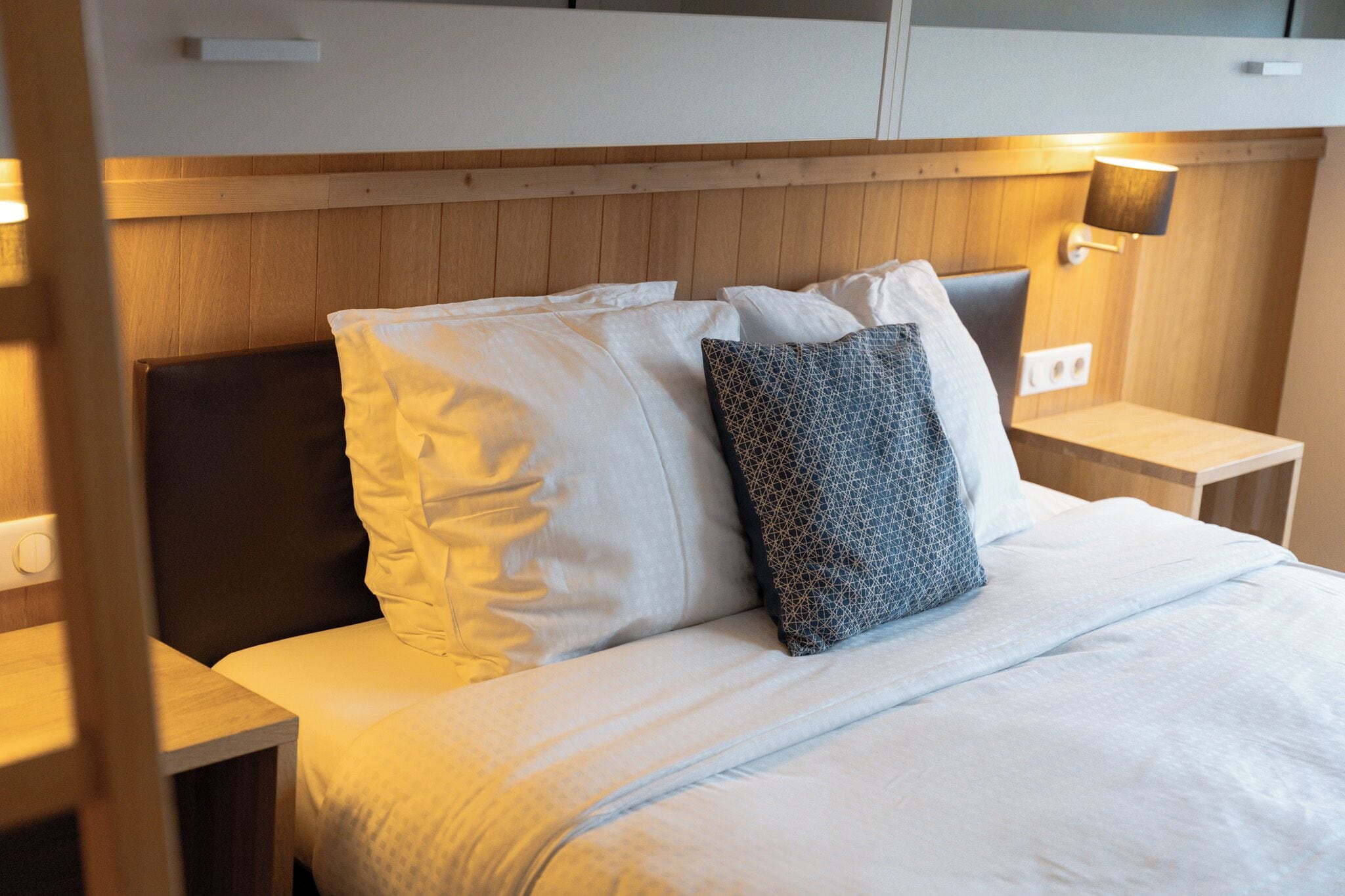 A neatly made bed with white and patterned pillows, wooden headboard, bedside tables with lamps, and a wooden panel wall.