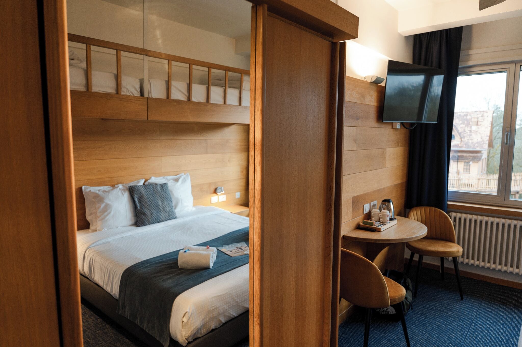 A hotel room with a bunk bed, a desk with chairs, a television, and a window with a view.