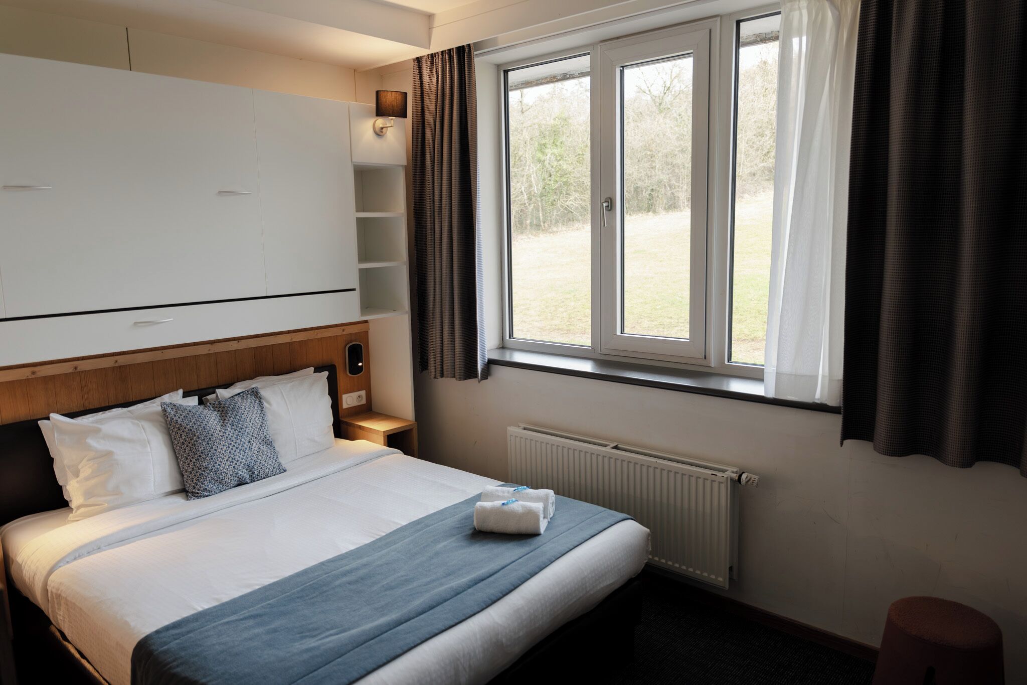 Una camera d'albergo con un letto, una finestra con tende, un termosifone e un armadio.