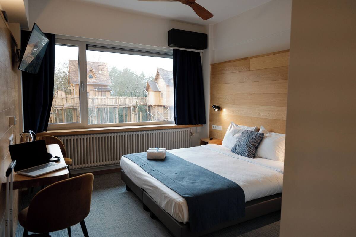 Une chambre d’hôtel avec un lit, un bureau avec un ordinateur portable, une chaise, une fenêtre avec des rideaux, un radiateur et une tête de lit en bois.