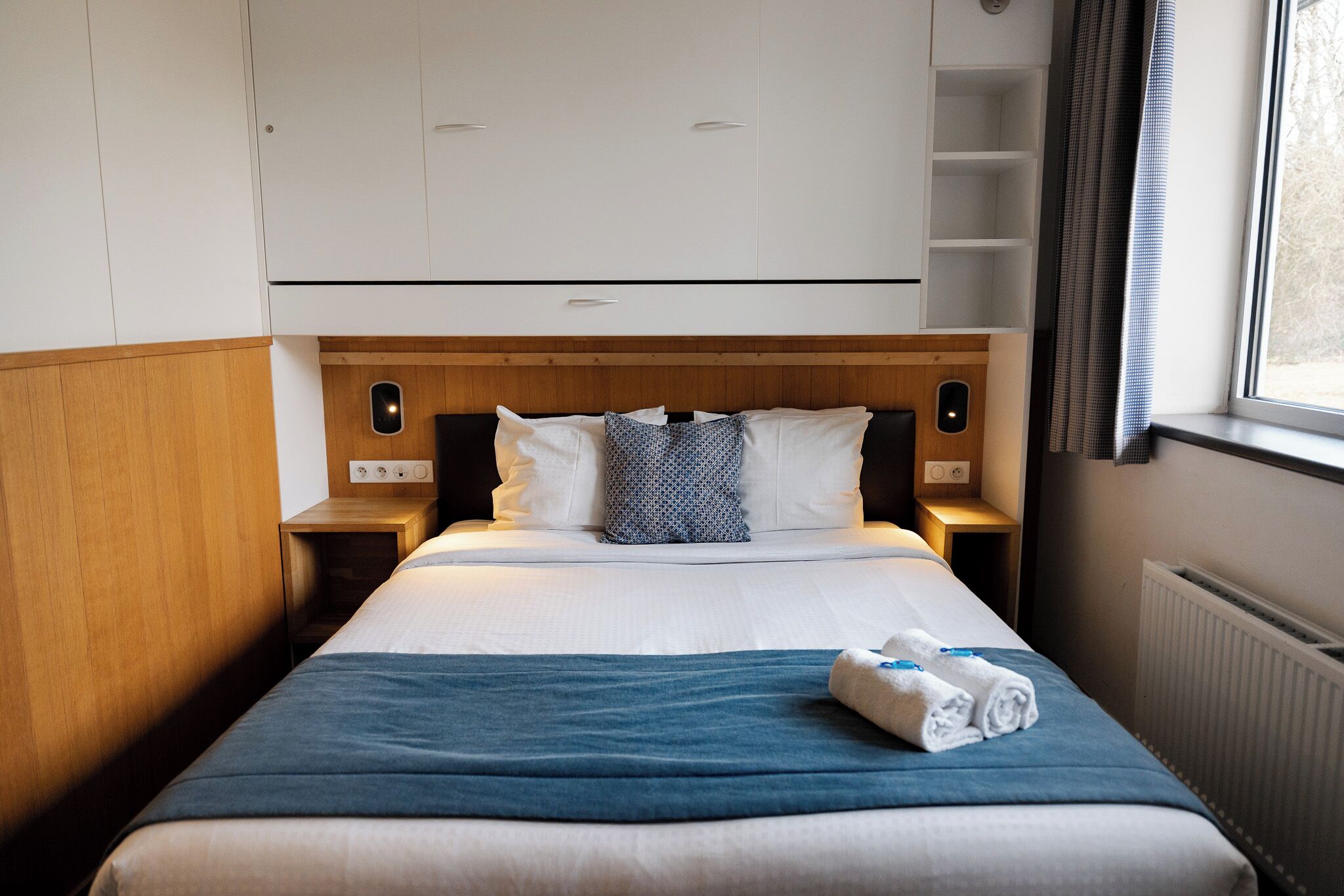 Ett hotellrum med en säng, ett träfärgat sänggavel, vita sängkläder, en blå filt och ett fönster med gardiner.