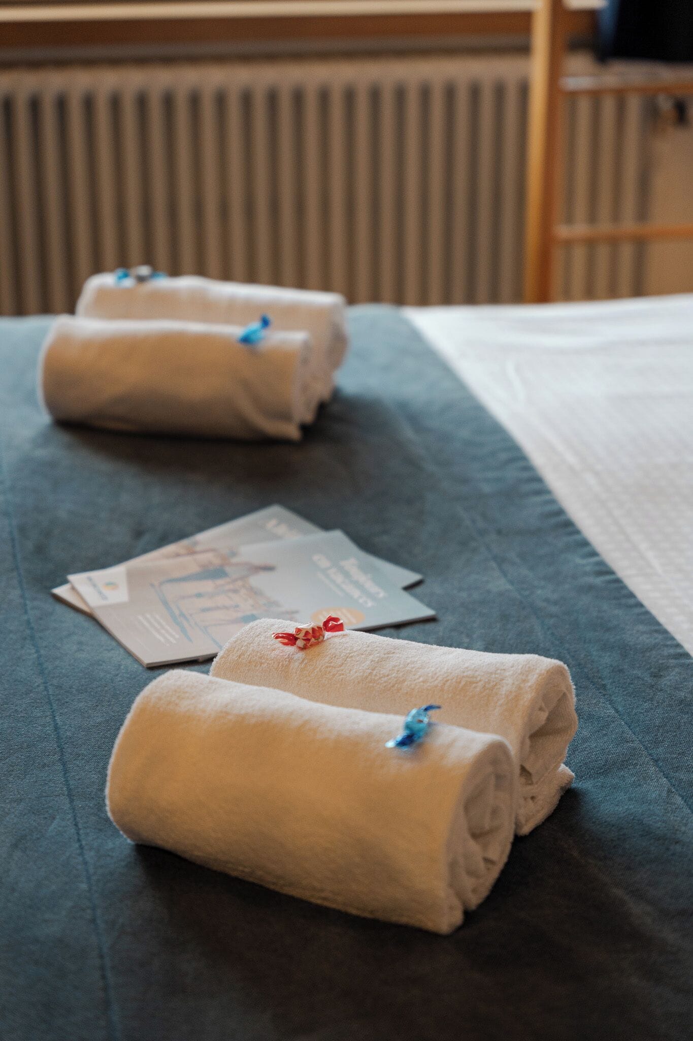 Un lit bien fait, avec des serviettes blanches roulées et une couverture bleue, une carte pliée et un petit objet décoratif rouge et blanc posé sur le lit.